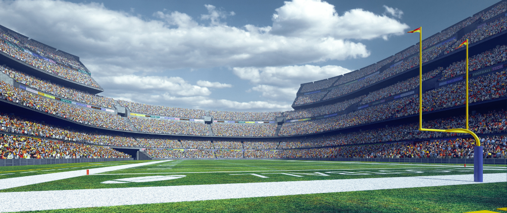 Full football stadium packed with cheering fans, bright green end zone turf, and yellow goalpost under a blue sky with white clouds.