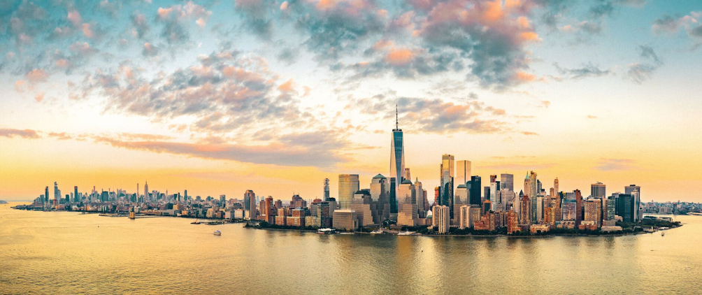 Panoramic view of Lower Manhattan, New York City at sunset, featuring the One World Trade Center skyscraper and surrounding skyline reflecting in the Hudson River.