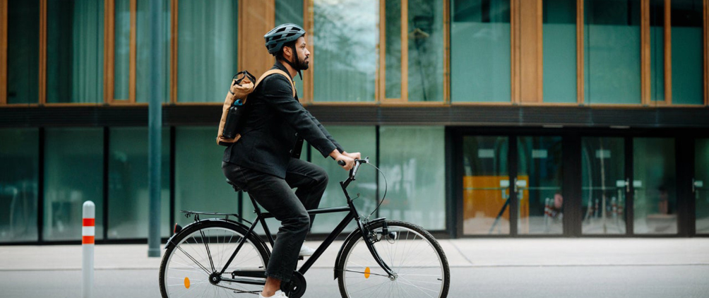 Urban commuter wearing a bicycle helmet and backpack riding a black city bike past a modern glass office building on a city street during daytime.