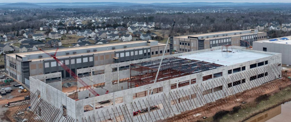 Aerial view of a large commercial warehouse construction site with steel framework, concrete walls, cranes, and workers, surrounded by a suburban neighborhood and rolling hills in the background.