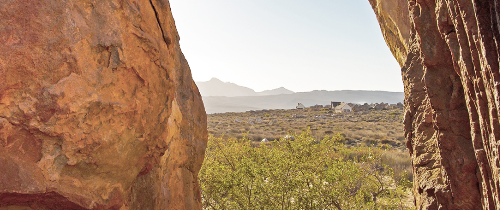 Desert landscape framed by two large reddish rock formations, overlooking a dry valley with scattered shrubs, distant mountains, and a small rural settlement under a clear sky