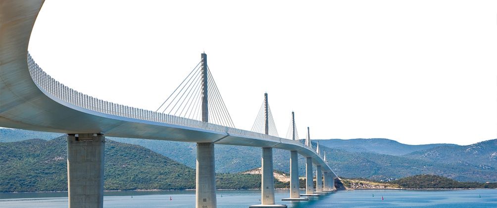Cable-stayed bridge curving across blue water with mountains in the background.