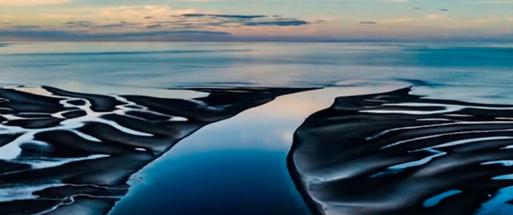 Aerial view of winding tidal channels flowing through dark coastal sand toward the horizon.