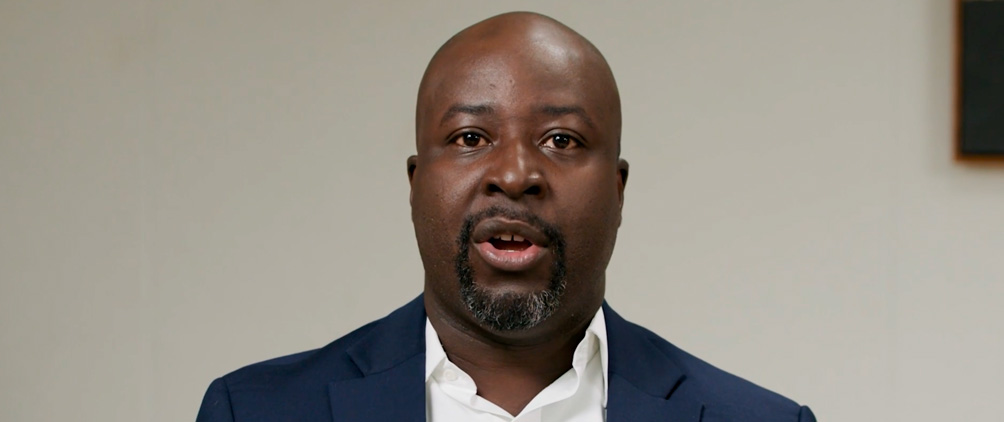 Man in a navy blazer speaking against a neutral background.