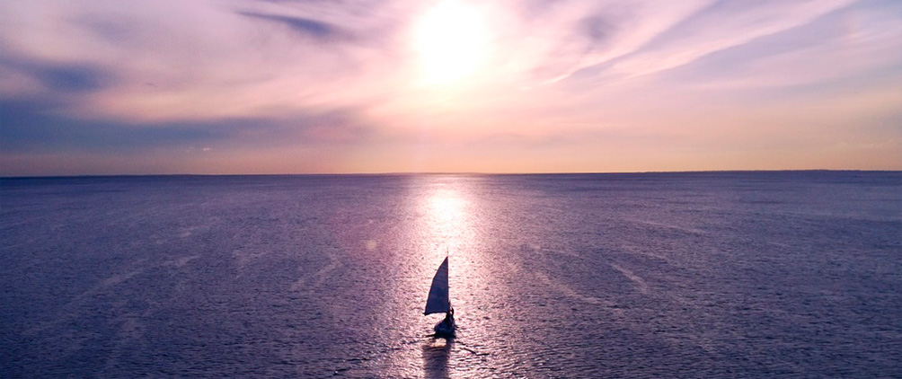 Sailboat crossing calm water at sunset beneath a pink and purple sky.