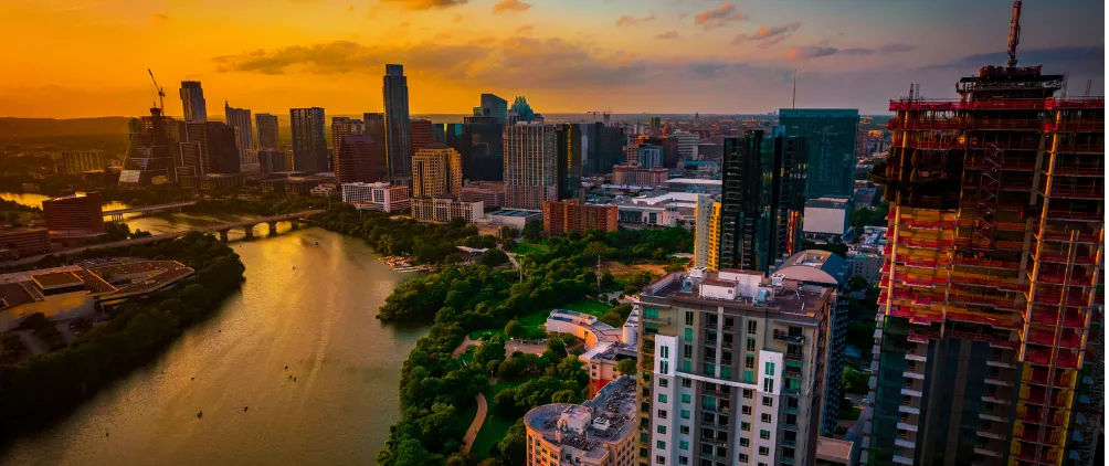 Aerial view of downtown Austin, Texas at sunset, with the Colorado River running through the city, modern high-rise buildings, and a skyscraper under construction in the foreground.