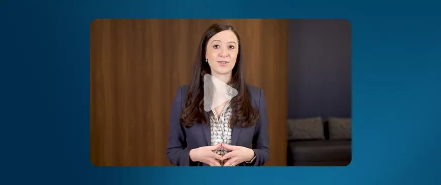 A woman with long dark hair, wearing a navy blazer over a patterned blouse, stands in front of a wooden wall and speaks while looking at the camera. A translucent play button overlay indicates this is a video still.