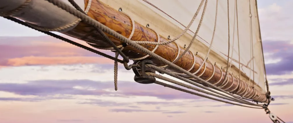 Close-up of a wooden boom with rigging and a sail attached, set against a pastel sky with soft pink and purple clouds at sunset.