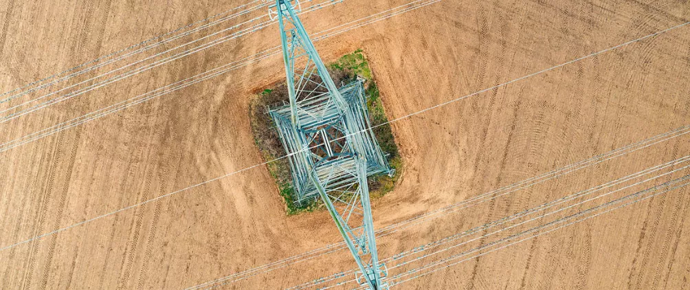 Aerial view of a high-voltage electricity pylon and power lines standing in the middle of a plowed agricultural field.