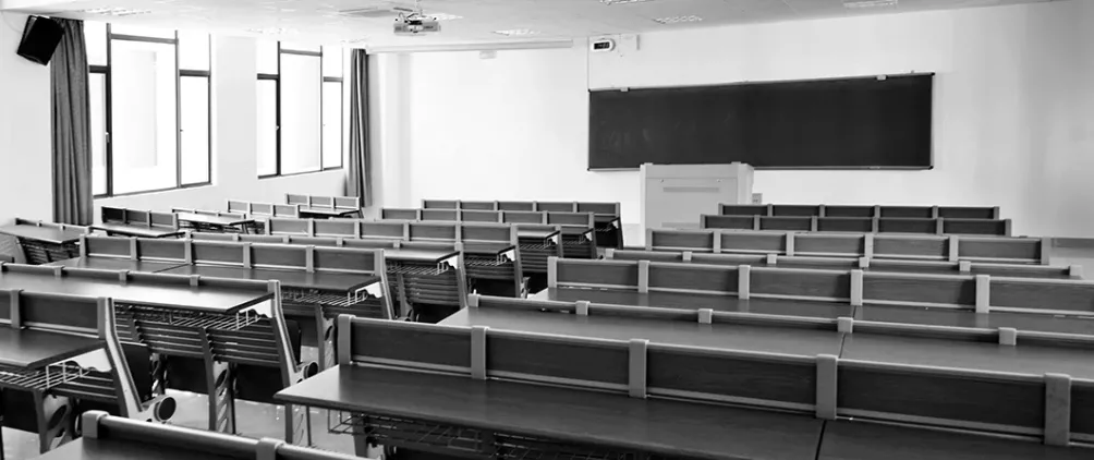 The image depicts an empty classroom or lecture hall. The room contains multiple rows of long desks with attached chairs, all facing toward the front. At the front of the room, there is a large chalkboard or blackboard mounted on the wall and a podium or lectern positioned in front of it. Several large windows on the left side let in natural light, and the room appears clean, orderly, and modern. The photo is in black and white, giving it a quiet, still atmosphere.