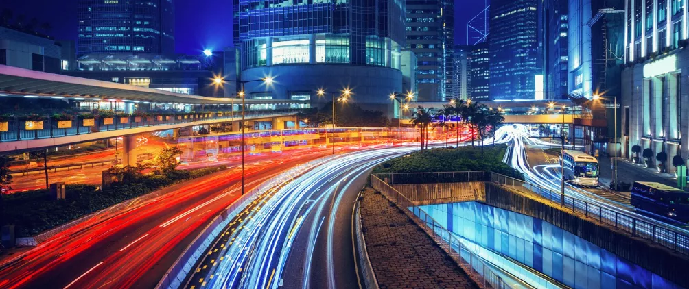 Nighttime urban cityscape with modern skyscrapers and elevated highways, featuring long-exposure light trails from fast-moving traffic, blue and red streaks curving through a futuristic downtown financial district.