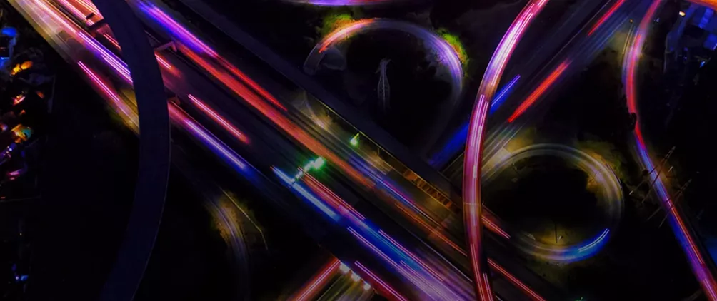 Aerial view of a highway interchange at night with colorful light trails representing leveraged finance, market complexity and the AI credit divide.